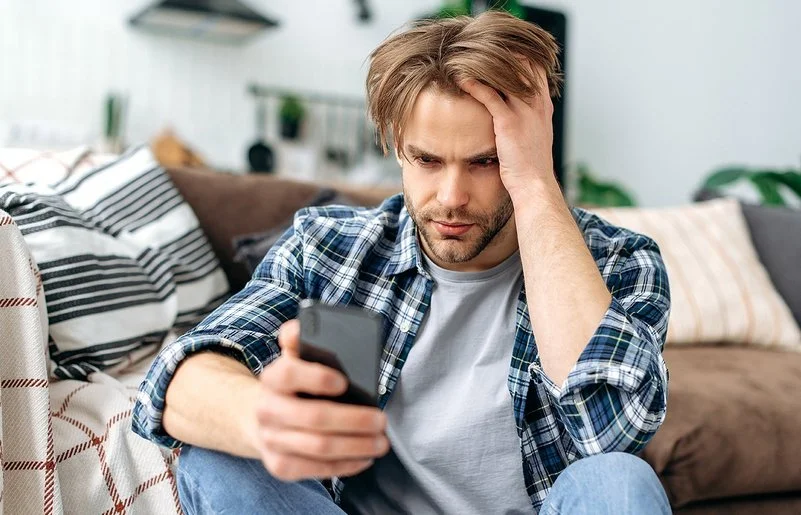 Ein junger Mann sitzt auf dem Fußboden vor einem Sofa. In der rechten Hand hält er ein Smartphone, mit der linken fährt er sich durch die Haare. Er schaut ernst auf das Display.