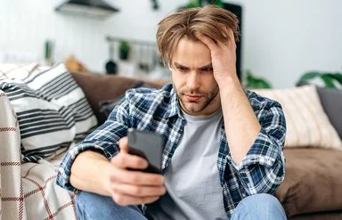 Ein junger Mann sitzt auf dem Fußboden vor einem Sofa. In der rechten Hand hält er ein Smartphone, mit der linken fährt er sich durch die Haare. Er schaut ernst auf das Display.