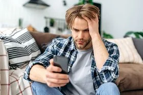 Ein junger Mann sitzt auf dem Fußboden vor einem Sofa. In der rechten Hand hält er ein Smartphone, mit der linken fährt er sich durch die Haare. Er schaut ernst auf das Display.