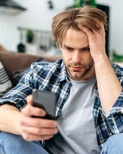 Ein junger Mann sitzt auf dem Fußboden vor einem Sofa. In der rechten Hand hält er ein Smartphone, mit der linken fährt er sich durch die Haare. Er schaut ernst auf das Display.