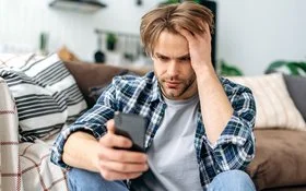 Ein junger Mann sitzt auf dem Fußboden vor einem Sofa. In der rechten Hand hält er ein Smartphone, mit der linken fährt er sich durch die Haare. Er schaut ernst auf das Display.