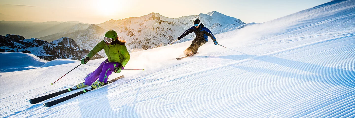 Ein Mann und eine Frau fahren auf Skiern rasant eine Piste hinab. Im Hintergrund sind schneebedeckte Berge zu sehen.