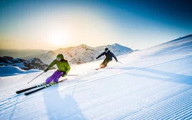 Ein Mann und eine Frau fahren auf Skiern rasant eine Piste hinab. Im Hintergrund sind schneebedeckte Berge zu sehen.