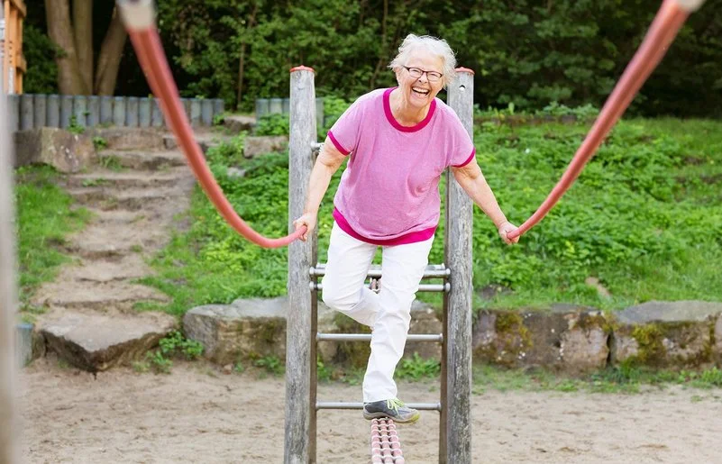 Eine grauhaarige Frau in Freizeitkleidung balanciert lachend über ein Klettergerüst auf einem Spielplatz.