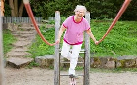 Eine grauhaarige Frau in Freizeitkleidung balanciert lachend über ein Klettergerüst auf einem Spielplatz.