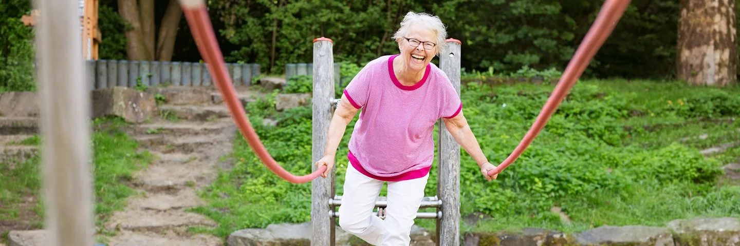 Eine grauhaarige Frau in Freizeitkleidung balanciert lachend über ein Klettergerüst auf einem Spielplatz.