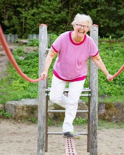 Eine grauhaarige Frau in Freizeitkleidung balanciert lachend über ein Klettergerüst auf einem Spielplatz.