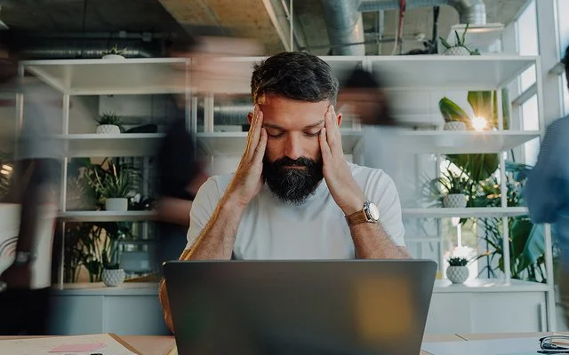Ein Mann sitzt am Schreibtisch vor einem Laptop. Er stützt die Ellenbogen auf die Tischplatte und fasst sich mit den Händen an die Schläfen. Seine Augen sind geschlossen.