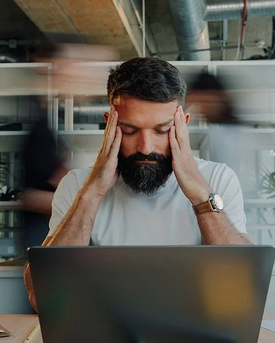 Ein Mann sitzt am Schreibtisch vor einem Laptop. Er stützt die Ellenbogen auf die Tischplatte und fasst sich mit den Händen an die Schläfen. Seine Augen sind geschlossen.