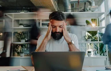 Ein Mann sitzt am Schreibtisch vor einem Laptop. Er stützt die Ellenbogen auf die Tischplatte und fasst sich mit den Händen an die Schläfen. Seine Augen sind geschlossen.