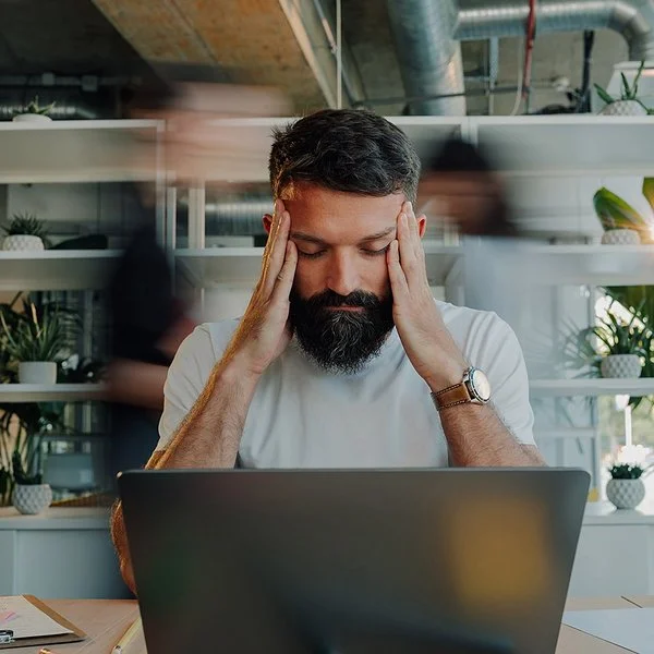 Ein Mann sitzt am Schreibtisch vor einem Laptop. Er stützt die Ellenbogen auf die Tischplatte und fasst sich mit den Händen an die Schläfen. Seine Augen sind geschlossen.