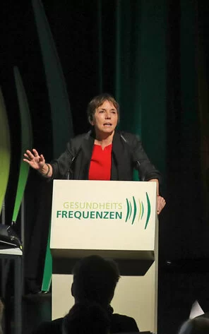 Eine Frau steht an einem Rednerpult. Im Hintergrund befindet sich ein farbenfrohes Bild.