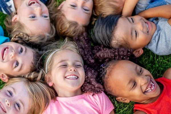 Mehrere Kinder liegen mit den Köpfen zusammen im Kreis auf einer Wiese und lächeln in die Kamera.