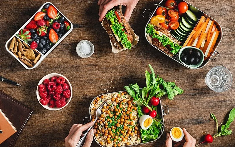 Auf einem Holztisch stehen mehrere Edelstahl-Lunchboxen, die mit frischem Gemüse, Obst und Vollkornprodukten gefüllt sind. In einer Dose liegen Kichererbsen auf Getreide, daneben halbierte Eier und Blattsalat. Eine weitere Dose enthält Gemüsesticks wie Karotten, Gurken und Tomaten. In einer dritten Dose befinden sich Beeren und Cracker. Zwei Hände greifen nach dem Essen, eine hält eine Gabel, die andere ein belegtes Sandwich. Frische Himbeeren und ein kleiner Behälter mit Salz stehen ebenfalls auf dem Tisch.