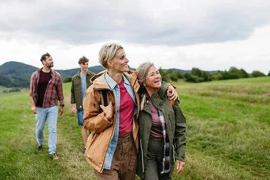 Eine reife Tochter hält ihre Mutter im Arm und wandert mit ihr und weiteren Familienmitgliedern durch die Natur.