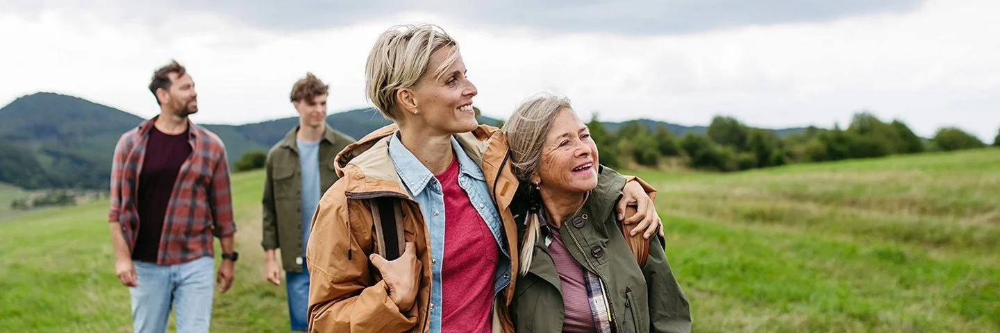 Eine reife Tochter hält ihre Mutter im Arm und wandert mit ihr und weiteren Familienmitgliedern durch die Natur.
