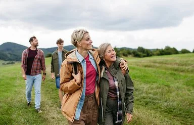 Eine reife Tochter hält ihre Mutter im Arm und wandert mit ihr und weiteren Familienmitgliedern durch die Natur.