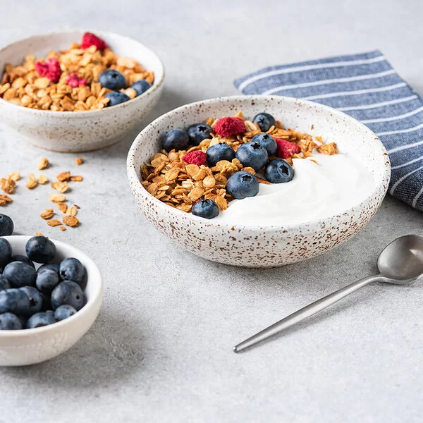 Nahaufnahme von Granola-Joghurtschalen mit Heidelbeeren und getrockneten Himbeeren auf einem grauen Betontisch.