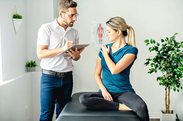 Ein Osteopath steht einer Frau gegenüber und macht Notizen auf einem Klemmbrett. Die Frau sitzt auf einer Liege und deutet mit ihrer linken Hand auf ihre rechte Schulter.