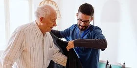 Ein männlicher Pfleger hilft einem alten Mann in eine Strickjacke. Beide Personen befinden sich in einem Wohnzimmer. Im Hintergrund ist ein Esstisch mit einer Sitzbank und mehreren Stühlen zu sehen.
