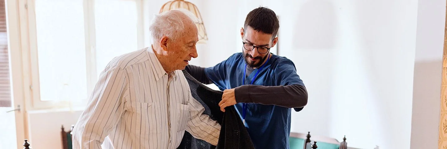 Ein männlicher Pfleger hilft einem alten Mann in eine Strickjacke. Beide Personen befinden sich in einem Wohnzimmer. Im Hintergrund ist ein Esstisch mit einer Sitzbank und mehreren Stühlen zu sehen.