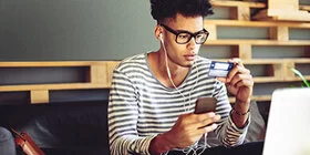 Ein junger Mann blickt prüfend auf seine Krankenversicherungskarte, die er in der linken Hand hält. In der rechten Hand hält er ein Smartphone.