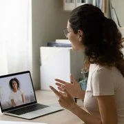 Eine junge Frau mit dunklen, lockigen Haaren und Brille, im beigen T-Shirt, sitzt am Schreibtisch und unterhält sich per Video-Chat mit einer anderen Frau.