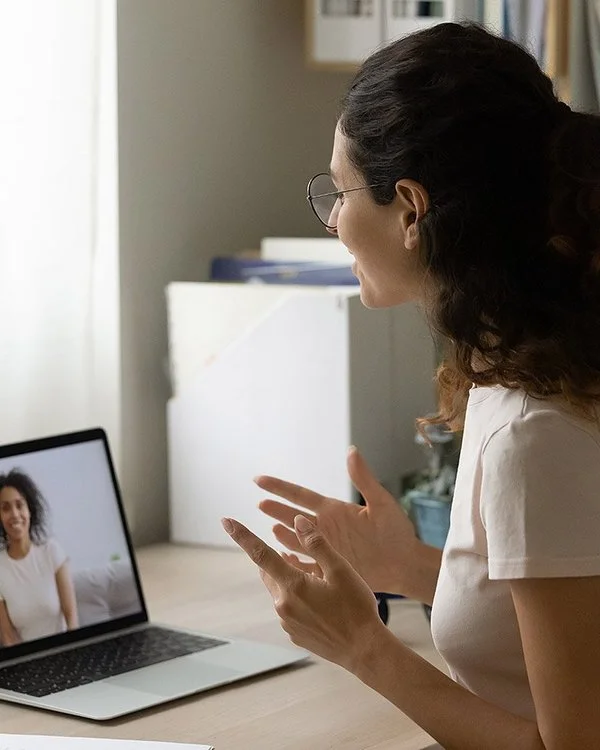 Eine junge Frau mit dunklen, lockigen Haaren und Brille, im beigen T-Shirt, sitzt am Schreibtisch und unterhält sich per Video-Chat mit einer anderen Frau.