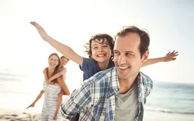 Eine Familie ist am Strand und genießt die Sonne. Die Eltern tragen die Kinder auf dem Rücken.