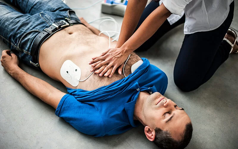 Ein Mann liegt am Boden und wird mittels Herzdruckmassage und Defibrillator reanimiert.