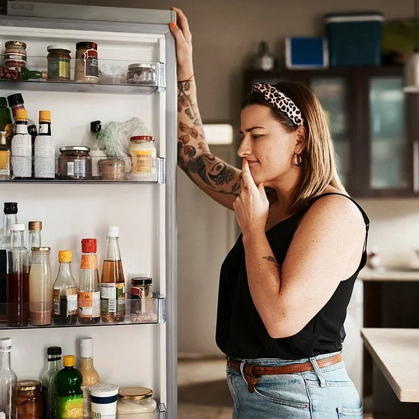 Eine Frau blickt unschlüssig in einen geöffneten Kühlschrank.