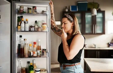 Eine Frau blickt unschlüssig in einen geöffneten Kühlschrank.