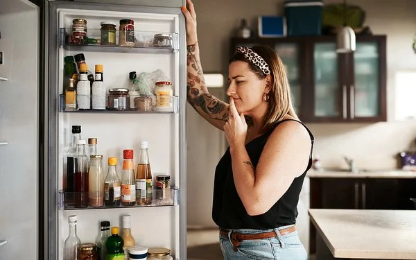 Eine Frau blickt unschlüssig in einen geöffneten Kühlschrank.