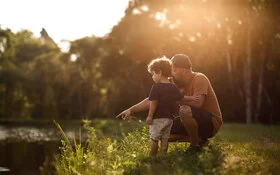 Ein Vater kniet mit seinem Sohn an einem Ufer und deutet aufs Wasser.