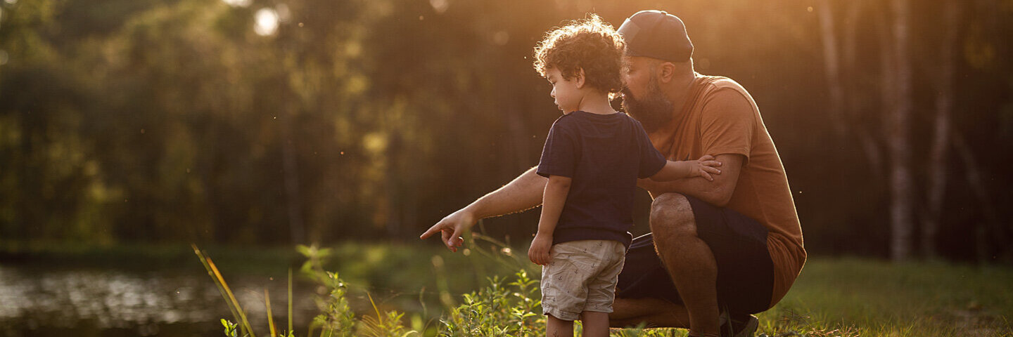 Ein Vater kniet mit seinem Sohn an einem Ufer und deutet aufs Wasser.