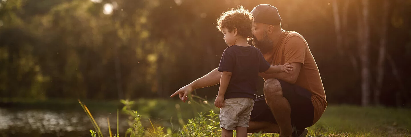 Ein Vater kniet mit seinem Sohn an einem Ufer und deutet aufs Wasser.