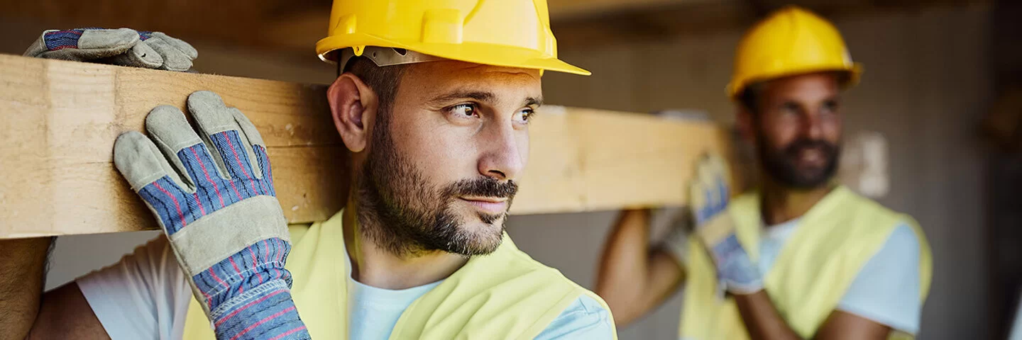 Zwei Bauarbeiter mit gelben Schutzhelmen und Warnwesten tragen gemeinsam einen Holzbalken durch eine Baustelle. Der vordere Mann blickt konzentriert nach links, beide tragen Arbeitshandschuhe. Im Hintergrund ist eine unfertige Innenkonstruktion zu sehen.