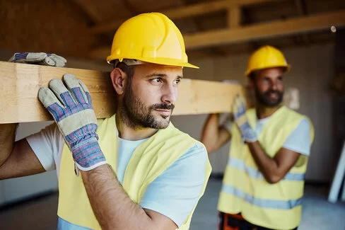 Zwei Bauarbeiter mit gelben Schutzhelmen und Warnwesten tragen gemeinsam einen Holzbalken durch eine Baustelle. Der vordere Mann blickt konzentriert nach links, beide tragen Arbeitshandschuhe. Im Hintergrund ist eine unfertige Innenkonstruktion zu sehen.