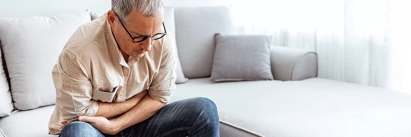 Ein älterer Mann sitzt zusammengekrümmt auf einem Sofa und fasst sich vor Schmerzen an den Bauch.