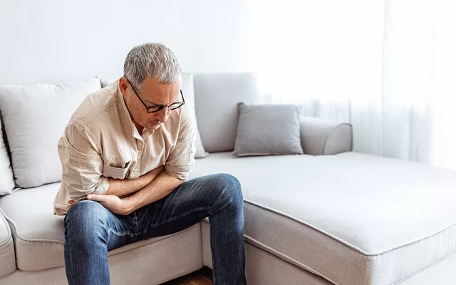 Ein älterer Mann sitzt zusammengekrümmt auf einem Sofa und fasst sich vor Schmerzen an den Bauch.