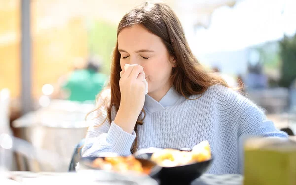 Eine junge Frau sitzt in einem Restaurant und schnupft in ein Taschentuch. Die Schüsseln mit den Speisen sind nur verschwommen zu erkennen.