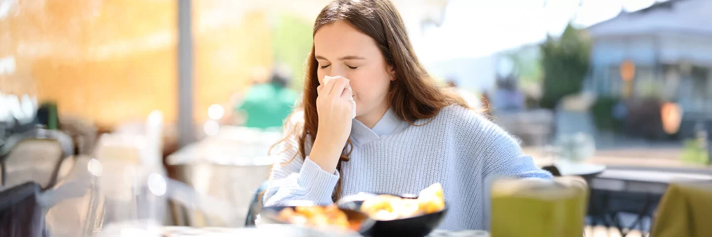 Eine junge Frau sitzt in einem Restaurant und schnupft in ein Taschentuch. Die Schüsseln mit den Speisen sind nur verschwommen zu erkennen.