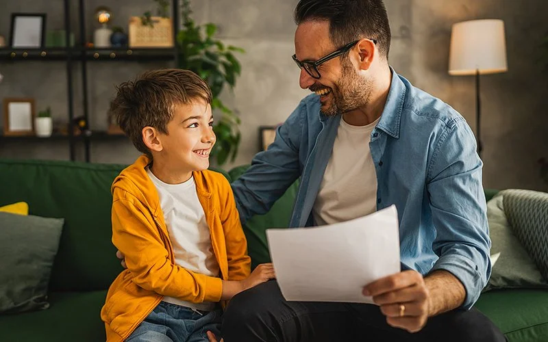 Ein Junge und sein Vater sitzen nebeneinander auf der Couch und schauen sich lächelnd an. Der Vater hält einen Test mit einer guten Note in der Hand.