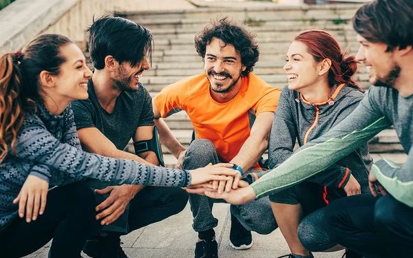 Eine Gruppe von fünf Personen in Joggingbekleidung – drei junge Männer und zwei junge Frauen – kniet im Halbkreis auf dem Absatz einer weitläufigen Außentreppe. Sie strecken ihre rechten Arme aus und legen ihre Hände in der Mitte des Halbkreises übereinander.