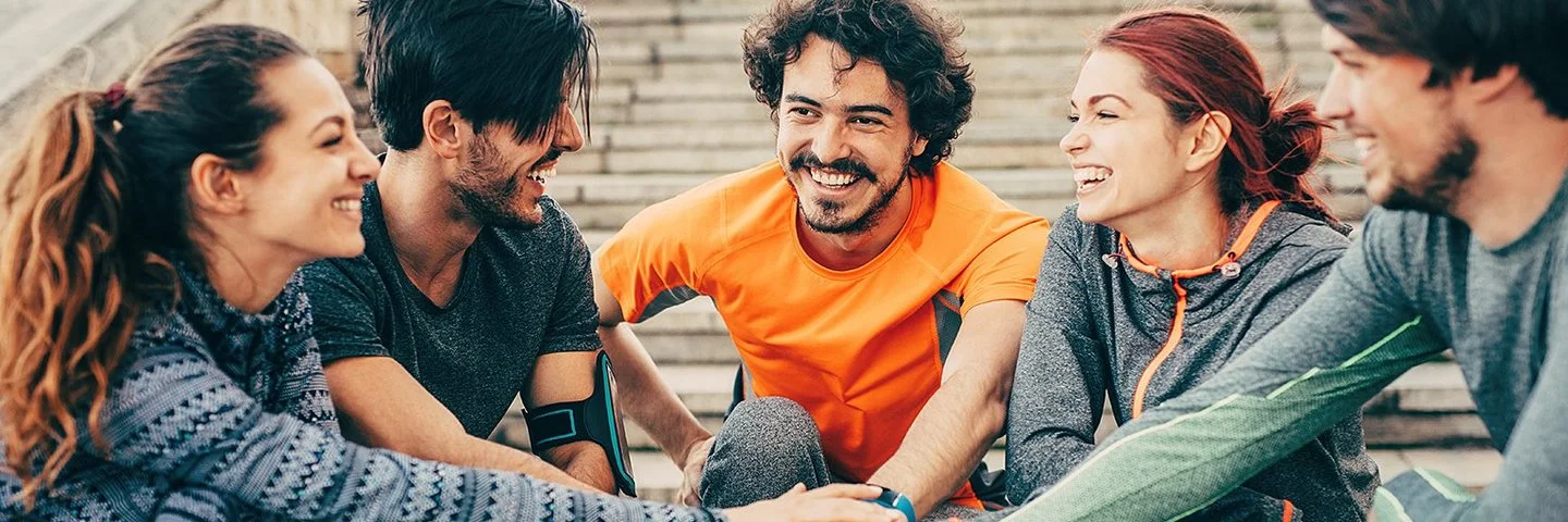 Eine Gruppe von fünf Personen in Joggingbekleidung – drei junge Männer und zwei junge Frauen – kniet im Halbkreis auf dem Absatz einer weitläufigen Außentreppe. Sie strecken ihre rechten Arme aus und legen ihre Hände in der Mitte des Halbkreises übereinander.