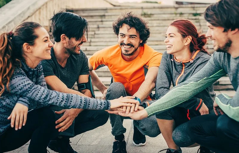Eine Gruppe von fünf Personen in Joggingbekleidung – drei junge Männer und zwei junge Frauen – kniet im Halbkreis auf dem Absatz einer weitläufigen Außentreppe. Sie strecken ihre rechten Arme aus und legen ihre Hände in der Mitte des Halbkreises übereinander.