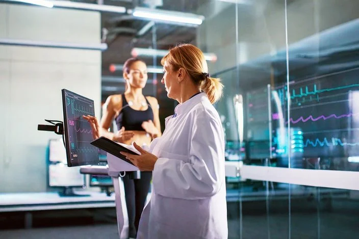 Sportwissenschaftler überprüft den EKG-Status einer Sportlerin, die im Hintergrund auf einem Laufband laufen auf einem Laufband mit Elektroden an ihrem Körper trainiert