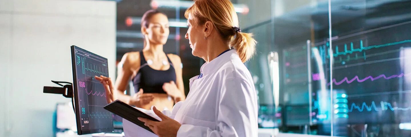 Sportwissenschaftler überprüft den EKG-Status einer Sportlerin, die im Hintergrund auf einem Laufband laufen auf einem Laufband mit Elektroden an ihrem Körper trainiert