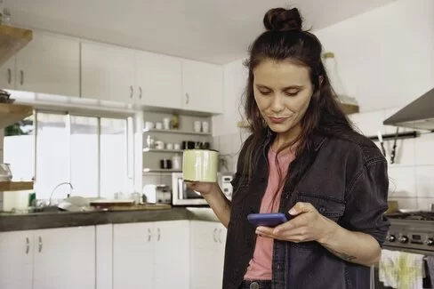 Junge Frau steht in der Küche mit einer Tasse Tee und schaut auf ihr Smartphone