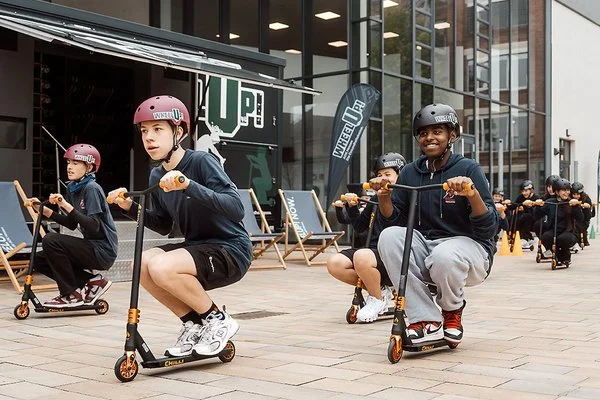 Eine Gruppe Kinder und Jugendlicher fährt mit Rollern auf einem Platz vor einem Gebäude.